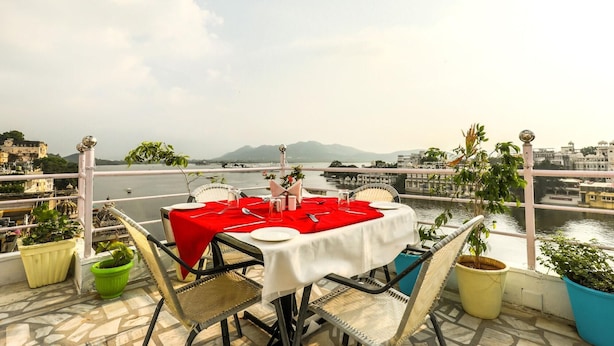 Una mesa para dos con mantel rojo, rodeada de sillas de mimbre, en un balcón con vistas a un río y edificios.