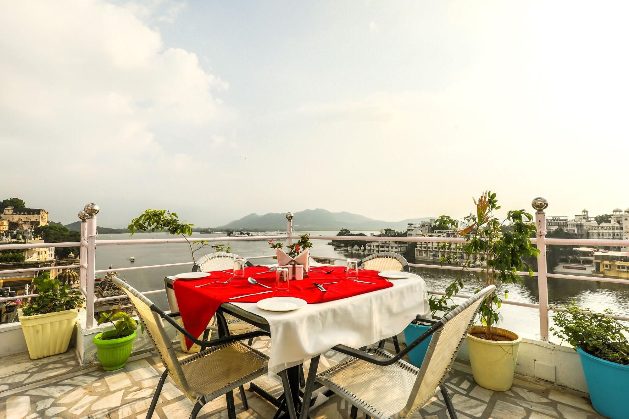 A table set for two with a red tablecloth, surrounded by wicker chairs, on a balcony with a view of a river and buildings.