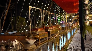 A buffet setup with food containers and serving utensils, surrounded by string lights and a reflective floor. - Mango Bloom River Resort (Ranikhet)