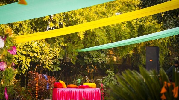 An outdoor area with a table set up for an event, surrounded by lush greenery and draped with colorful fabric.