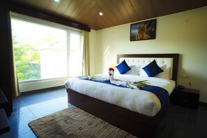 A bedroom with a large bed, a view of greenery outside, and a painting on the wall. - Mango Bloom River Resort (Ranikhet)