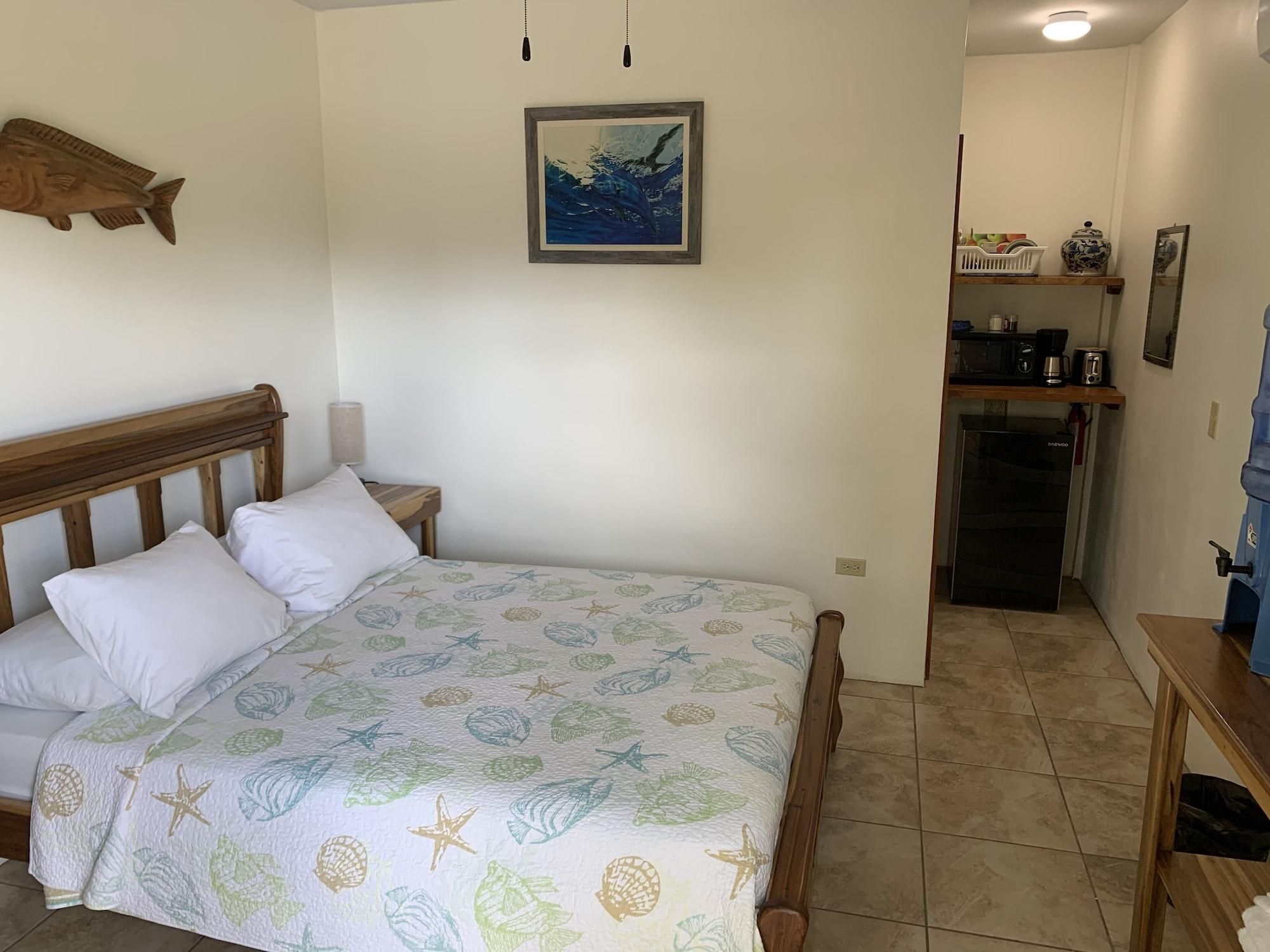 A bedroom with a bed, a wooden headboard, a wall-mounted fish decoration, a framed painting, and a kitchenette area with a refrigerator and coffee maker.