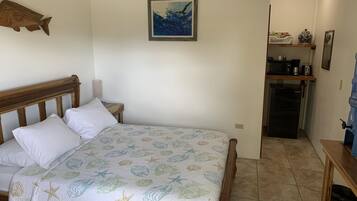 A bedroom with a bed, a wooden headboard, a wall-mounted fish decoration, a framed painting, and a kitchenette area with a refrigerator and coffee maker.