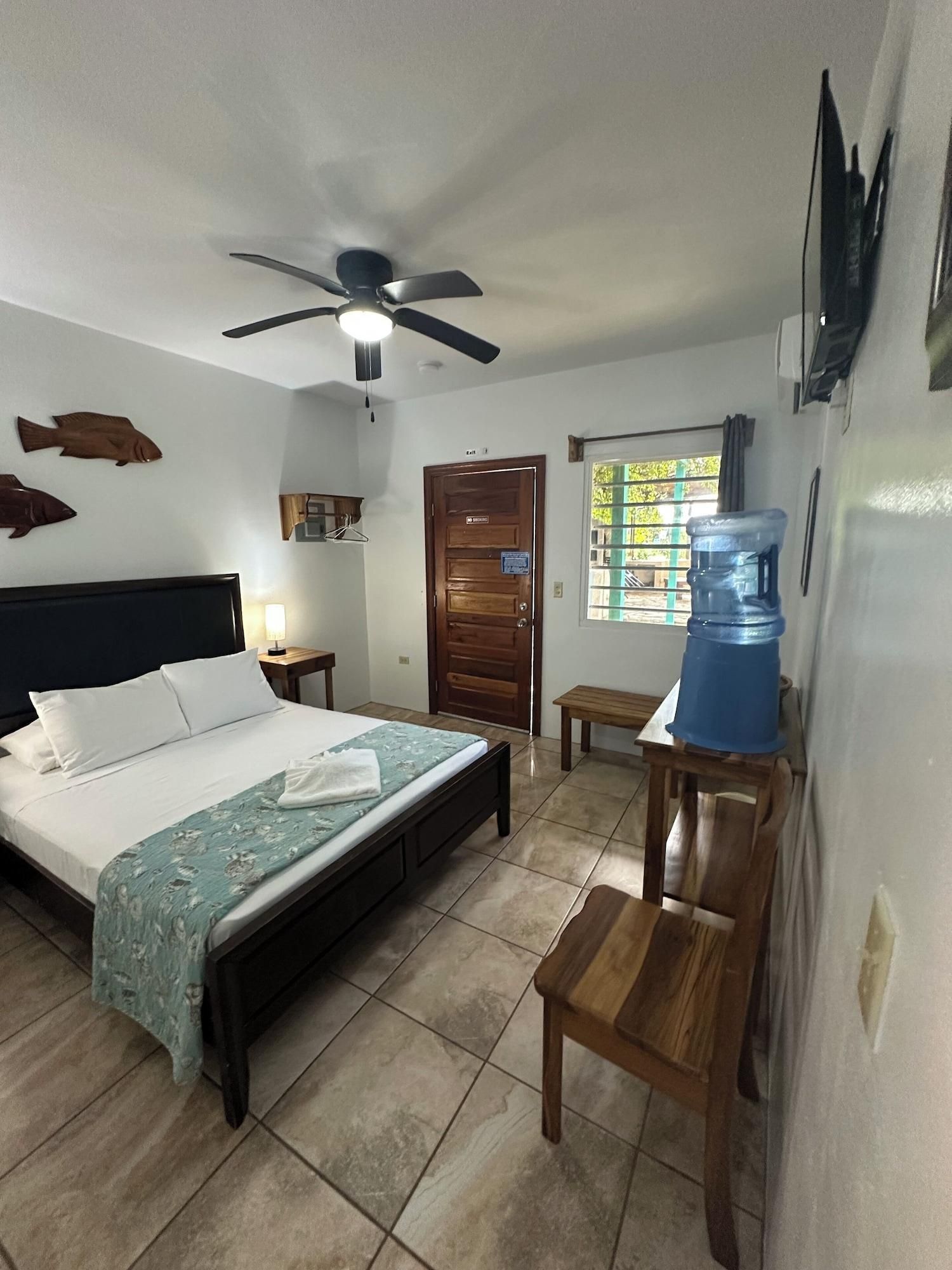 A bedroom with a bed, a ceiling fan, a wooden door, a window, and a water dispenser.
