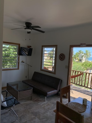 Una habitación con sofá, una mesa de vidrio, televisor y balcón con vistas a áreas verdes y un edificio.