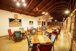 Un restaurant avec des tables et des chaises en bois, un plafond haut avec des poutres apparentes et des tableaux accrochés aux murs.