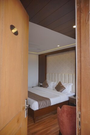 A hotel room with a wooden door, a bed with white bedding and brown pillows, a wooden headboard, and a red chair. - Sangam By Stotrak - Mussoorie (Dehradun)