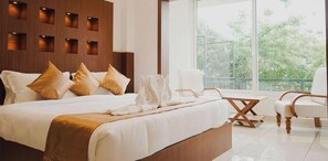 A modern hotel room with a large bed, a seating area with chairs, and a view of greenery through large windows.