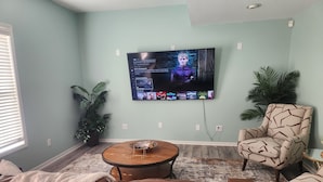 A living room with a television mounted on the wall, a brown leather couch, a patterned armchair, and a small table with a bowl. - CLOSE TO STADIUM! 2-bdroom lower level open-concept home (Kansas City)