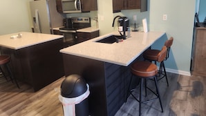 A kitchen with dark wooden cabinets, a stainless steel refrigerator, a microwave, a stove, and a sink. There are two bar stools and a trash can. - CLOSE TO STADIUM! 2-bdroom lower level open-concept home (Kansas City)