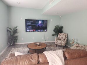 A living room with a flat-screen TV mounted on the wall, a brown leather sofa, a patterned armchair, a small round coffee table, and two potted plants. - CLOSE TO STADIUM! 2-bdroom lower level open-concept home (Kansas City)