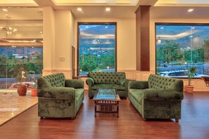 A spacious lobby with a green sofa set, a glass coffee table, and large windows offering a view of a parking area and greenery. - Katoch Grand By Vivaan (Manali)