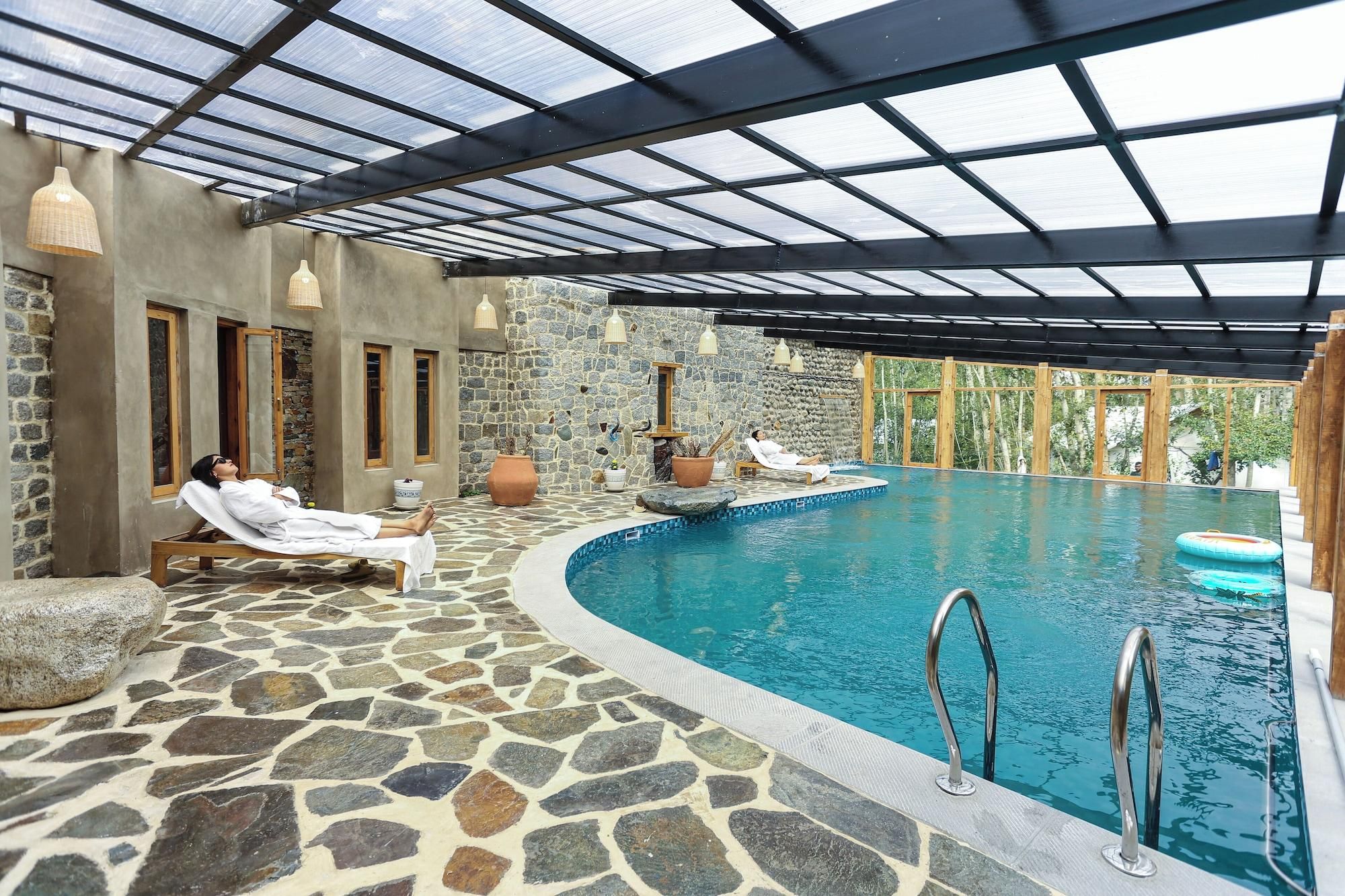 An indoor swimming pool with stone flooring, a glass ceiling, and a person relaxing by the pool.