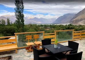 A terrace with wooden furniture, glass railings, and a view of mountains. - Stone Hedge Hotel Hunder (Leh)