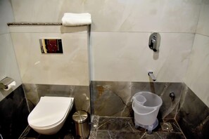 A bathroom with a toilet, a trash bin, a towel rack, and a shower area. - Hotel Aarna (Jaipur)