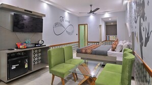 Una habitación moderna con una cama, sillas verdes, un televisor, un ventilador de techo y una pared con diseño decorativo.