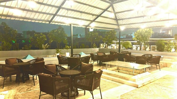 An outdoor seating area with wicker chairs and tables, a covered roof, and a view of the cityscape. - Hotel Natraz New Delhi (New Delhi)