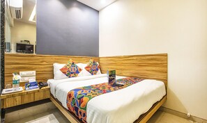 A hotel room with a wooden headboard, a bed with a colorful geometric-patterned bedspread, a bedside table with a phone, a water bottle, and a magazine. - Fabexpress Marol Metro (Mumbai)