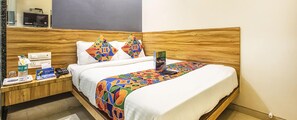A hotel room with a wooden headboard, a bed with a colorful geometric-patterned bedspread, a bedside table with a water bottle, a phone, and a magazine. - Fabexpress Marol Metro (Mumbai)
