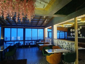 An indoor dining area with tables and chairs, large windows, and hanging floral decorations. - Hotel Grand Hill (Dalhousie)