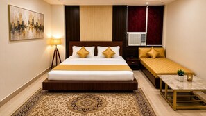 A hotel room with a large bed, bedside table, bedside lamp, and a small table with a plant. - Amaira Hotels And Banquets (Ghaziabad)