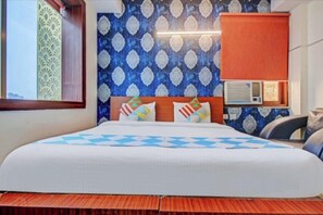 A hotel room with a bed, a headboard with decorative pillows, a wall-mounted air conditioning unit, and a window with a view. - Amaira Hotels And Banquets (Ghaziabad)