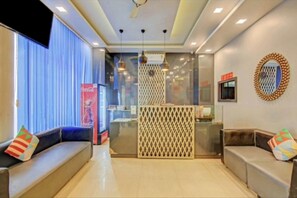 Un hall de hotel con mostrador de recepción, dos sofás con cojines de colores, un refrigerador Coca-Cola y un espejo decorativo en la pared.