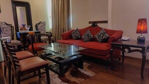 Uma sala de estar com um sofá vermelho, uma mesa de centro de vidro, cadeiras de madeira, uma mesinha com abajur e um espelho na parede.