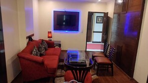 A living room with a red sofa, a glass coffee table, a TV mounted on the wall, and a door leading to another room. - Hotel Comfort Inn Bl (Haldwani)