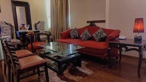 A living room with a red sofa, glass coffee table, wooden chairs, a small table with a lamp, and a mirror on the wall. - Hotel Comfort Inn Bl (Haldwani)