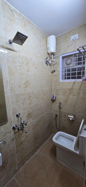 A bathroom with a shower, toilet, and a window with a fan. - Nj Residency (Chennai)