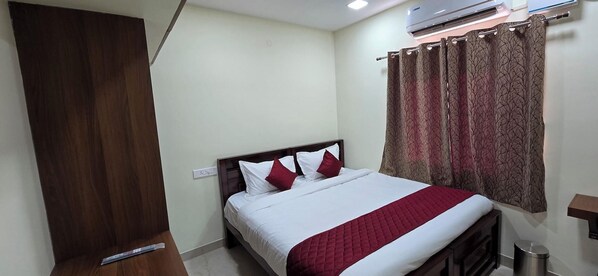 A hotel room with a bed, a wardrobe, an air conditioning unit, a curtain, and a trash bin.