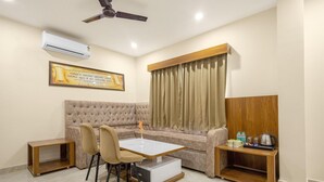 Ein Zimmer mit einer Couch, einem Esstisch mit Stühlen, einem Deckenventilator, einer Klimaanlage, einem Fenster mit Vorhängen und einem kleinen Holznachttisch mit Gegenständen darauf.