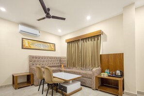 Una habitación con sofá, una mesa de comedor con sillas, ventilador de techo, equipo de aire acondicionado, ventana con cortinas y una pequeña mesita de madera con objetos encima.