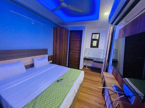 A hotel room with a bed, a desk with a TV, a ceiling fan, and a small bathroom. - Hotel Surya International (New Delhi)