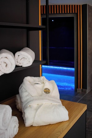 Camera d'albergo con un tavolo di legno su cui sono presenti asciugamani arrotolati e un accappatoio, con una piscina illuminata di blu sullo sfondo.