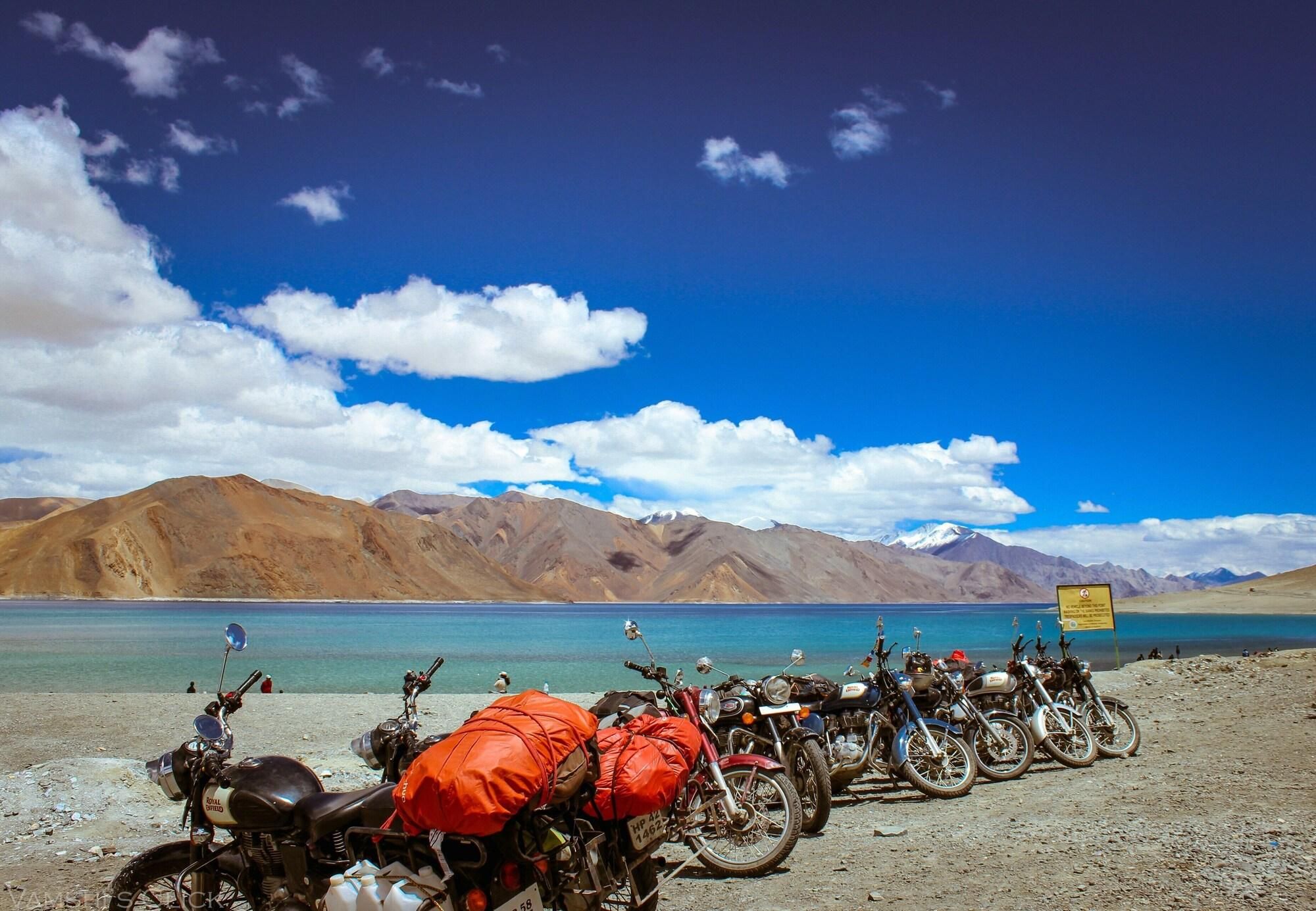 Motorcycles parked by a lake with mountains in the background.