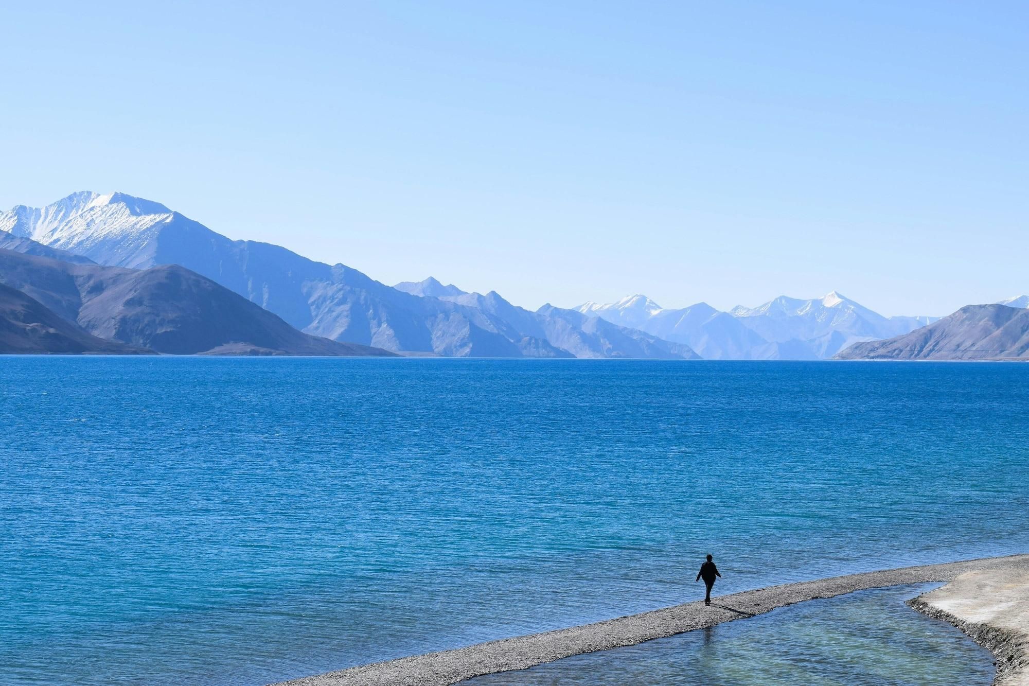 A person walking along a pebbly shore by a calm blue lake with mountains in the background.
