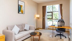 Ein Hotelzimmer mit einer Sitzecke, einem kleinen runden Tisch, einem Schreibtisch mit Stuhl, einer Lampe und einem fenster mit Vorhängen.