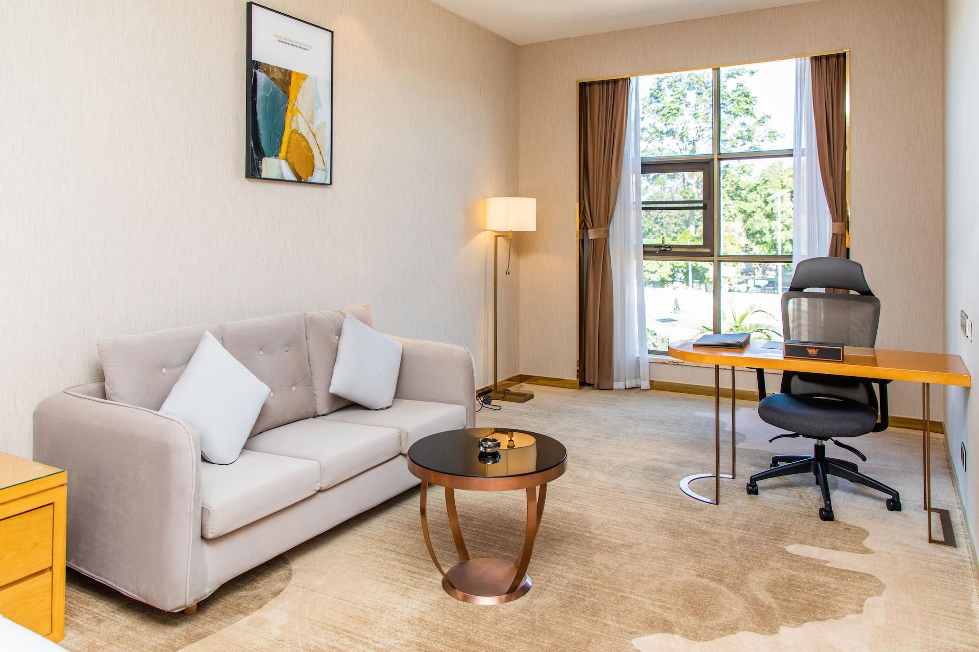 Habitación de hotel con sofá, una pequeña mesa redonda, escritorio con silla, lámpara y ventana con cortinas.