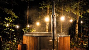Uma sauna de madeira ao ar livre com lareira visível, rodeada por árvores e luzes de Natal.