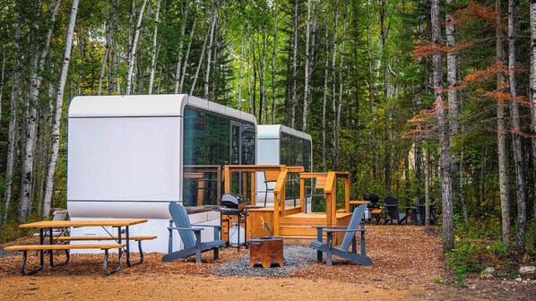 Een moderne cabin met een groot raam, houten terras en buitenzitplaatsen.