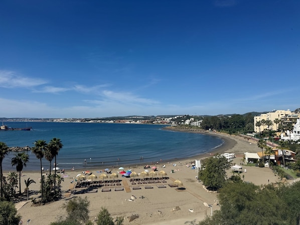 A beach with sun loungers, umbrellas, and a clear view of the sea and distant buildings. - Estepona Golf, Los Pintores (Estepona)