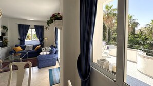 A living room with a sofa, a chair, a coffee table, and a view of a pool and palm trees. - Estepona Golf, Los Pintores (Estepona)