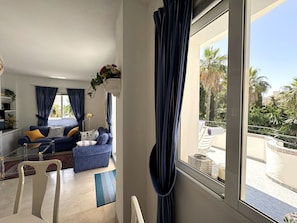 A living room with a sofa, a chair, a coffee table, and a view of a pool and palm trees. - Estepona Golf, Los Pintores (Estepona)