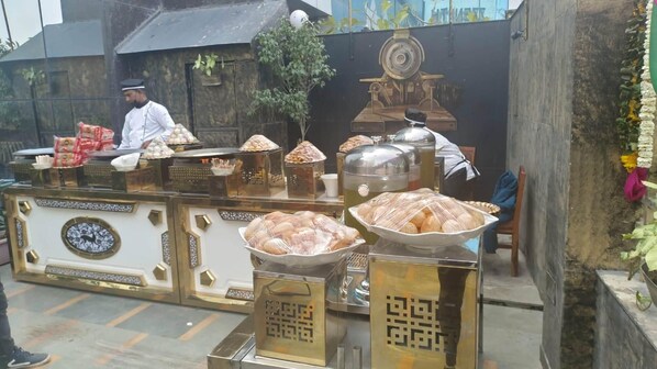 A food stall with various food items displayed in metal containers, a person in uniform behind the counter, and a decorative wall in the background. - Zinith Hotel (New Delhi)