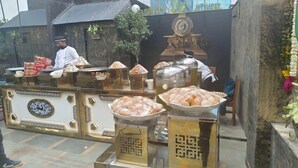 A food stall with various food items displayed in metal containers, a person in uniform behind the counter, and a decorative wall in the background. - Zinith Hotel (New Delhi)