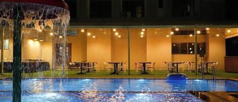 A pool with a water feature, tables, chairs, and a building with windows and lights.