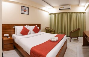 A hotel room with a bed, two chairs, a desk, an air conditioning unit, a painting on the wall, and a curtain. - Metropark Hotel (Mawal)