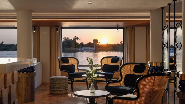 A room with a view of a sunset over water, featuring wicker chairs, a round table with a vase, and a bar area with a wooden counter. - Soleil Nile Cruise (Cairo)